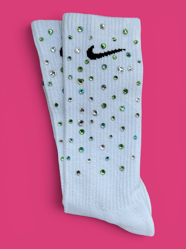 A pair of ribbed crew socks embellished with rhinestones and featuring a black embroidered Nike swoosh, displayed on a solid pink background.