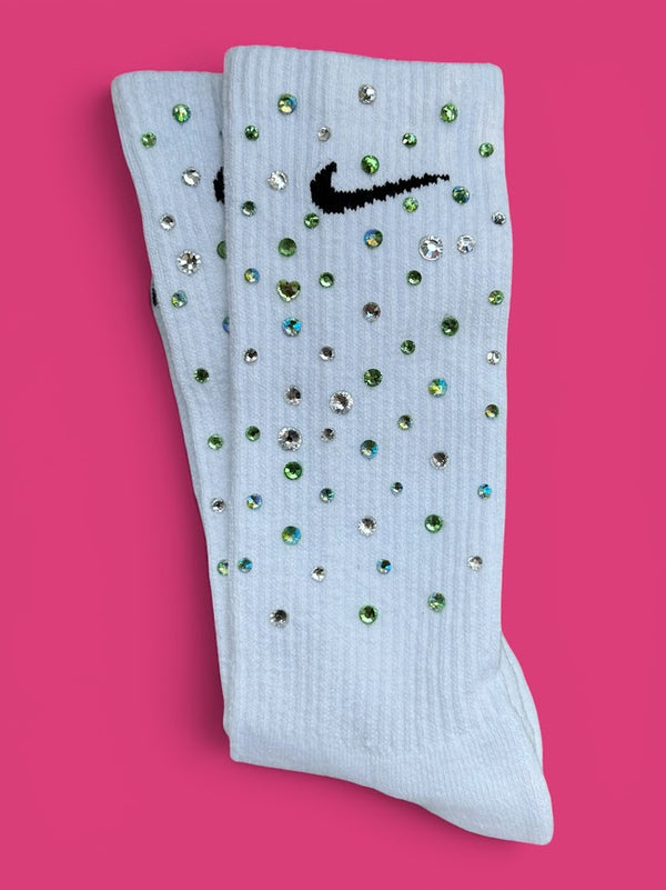 A pair of ribbed crew socks embellished with rhinestones and featuring a black embroidered Nike swoosh, displayed on a solid pink background.
