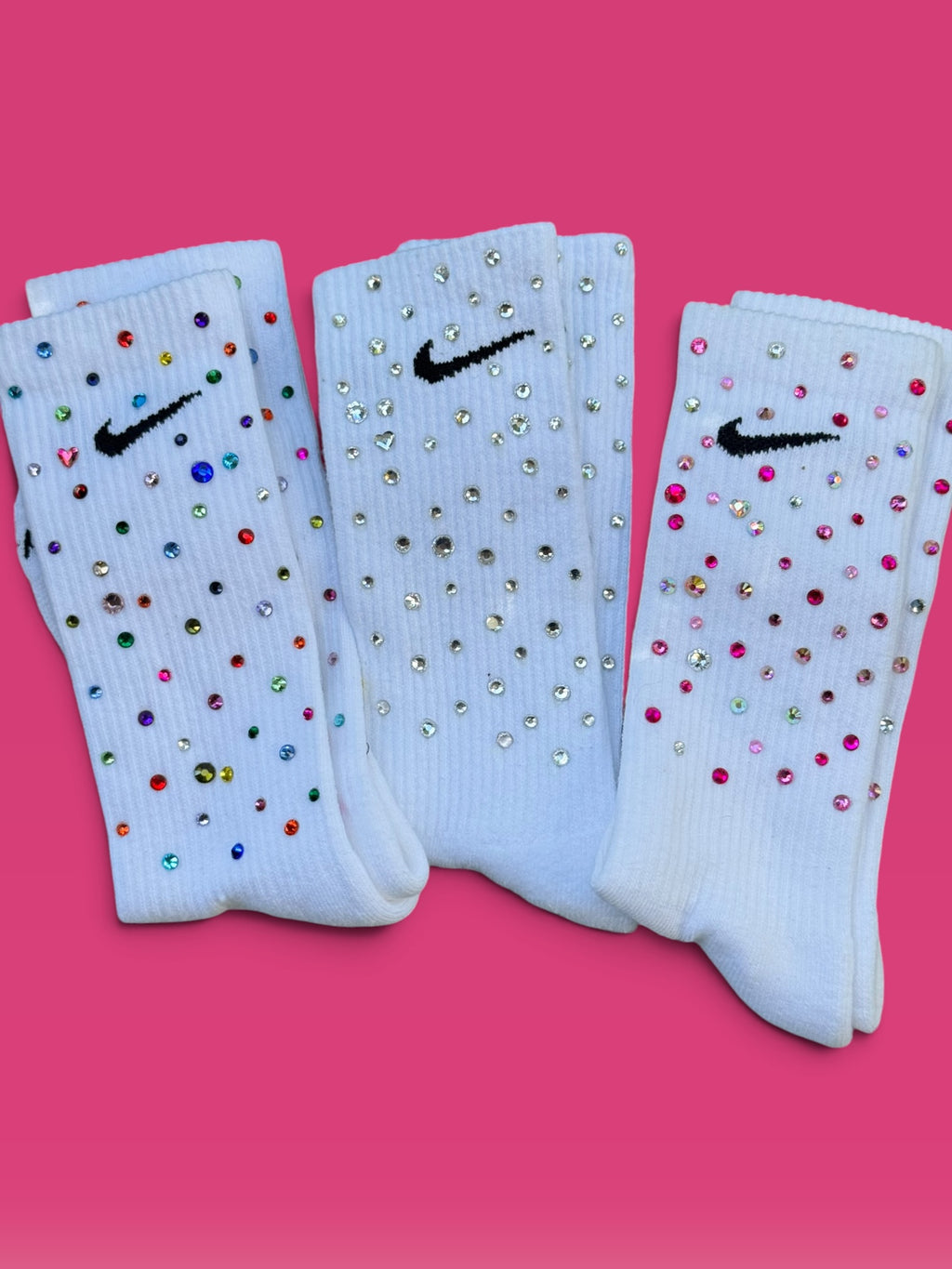 Three white Nike crew socks embellished with rhinestones, arranged side by side on a vibrant pink background.