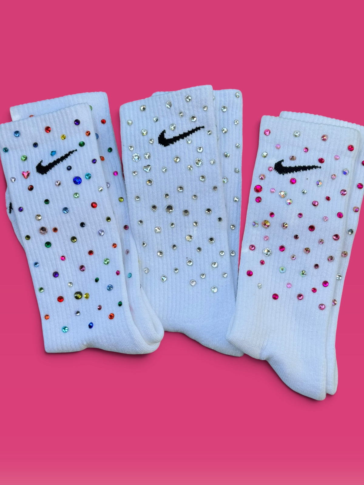 Three white Nike crew socks embellished with rhinestones, arranged side by side on a vibrant pink background.