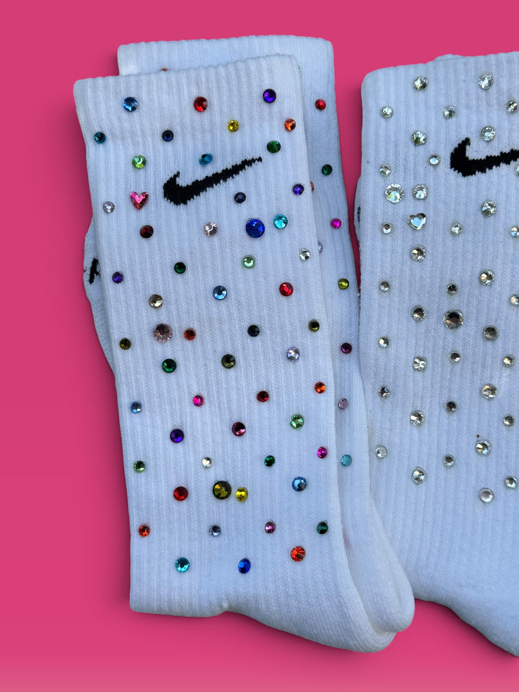 A pair of white Nike crew socks embellished with colorful rhinestones, displayed on a solid pink background.