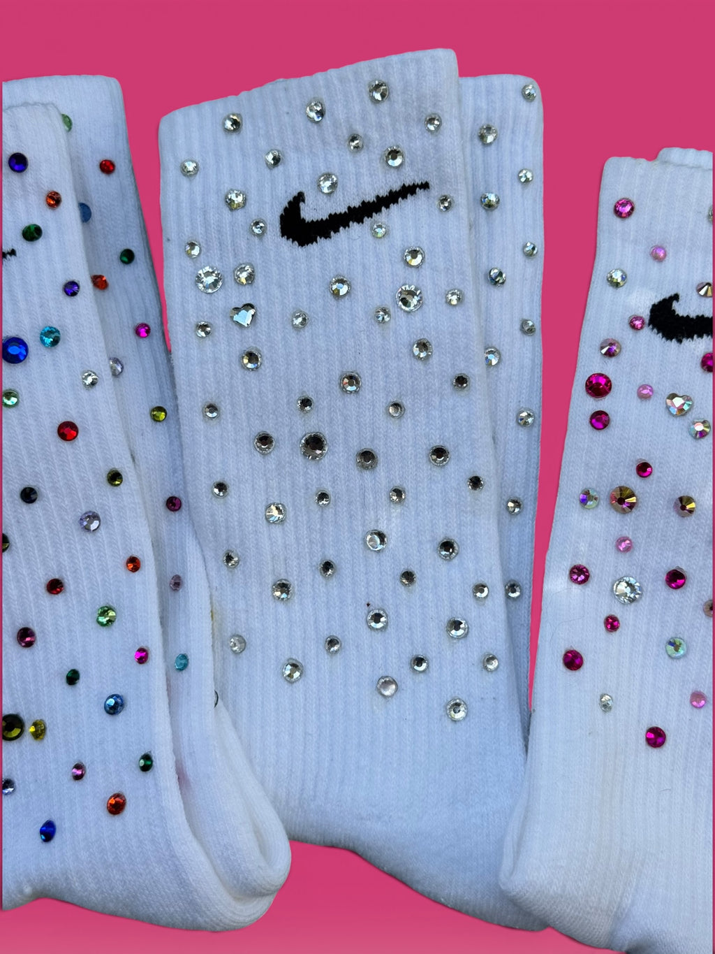 Three white Nike crew socks embellished with rhinestones and colorful gems, displayed against a solid pink background.