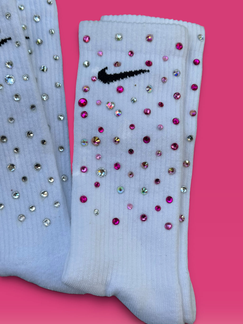 A pair of white Nike crew socks embellished with pink and silver rhinestones, displayed on a solid pink background.